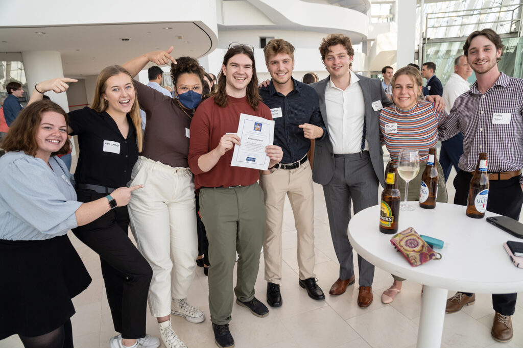 A group of seven people standing indoors, smiling and holding a certificate. Some have beverages and are casually dressed. They appear to be at a social or professional event, celebrating their business memberships.