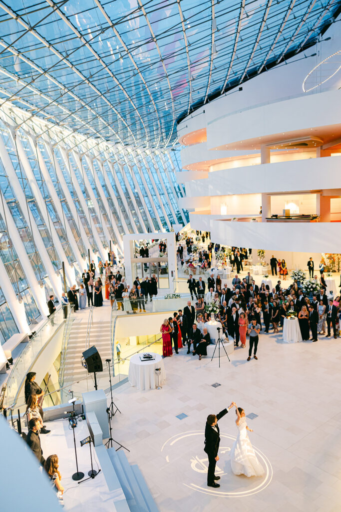 A couple dances on a spacious, modern indoor venue with a large, glass roof at the Kauffman Center. Many guests are gathered around, some seated and others standing, with tables and musical equipment visible—an ideal setting you can rent for your special occasion.