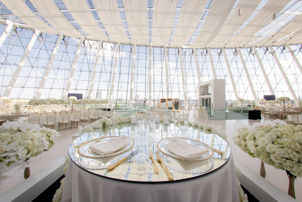 A luxurious, sunlit banquet hall set for a formal event with an elegantly decorated round table at the Rent Kauffman Center. The room features floor-to-ceiling windows and white floral arrangements.