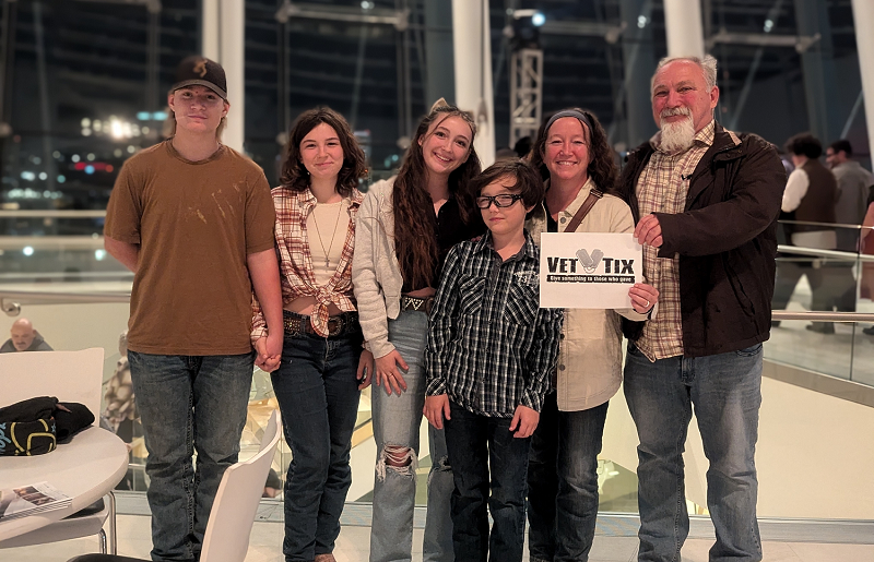 A group of six smiling patrons pose for a photo in a row in Brandmeyer Great Hall. Two patrons hold a small white paper sign that reads Vet Tix.