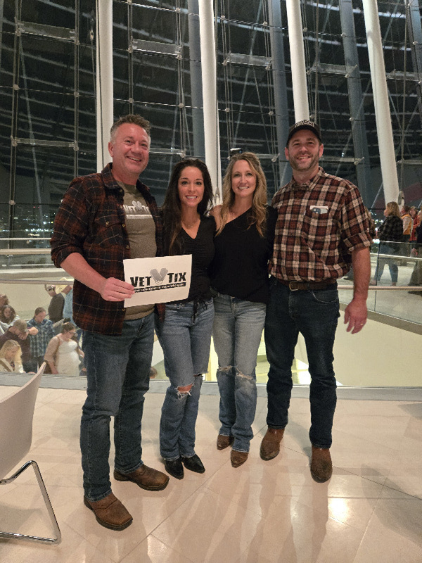 In the foreground, four standing patrons in jeans, flannel shirts and cowboy boots pose for a photo in Brandmeyer Great Hall. One patrons holds a white paper sign that reads "Vet Tix: Give Something for Those Who Gave." In the background, the white columns and glass panels of Brandmeyer Great Hall are visible.