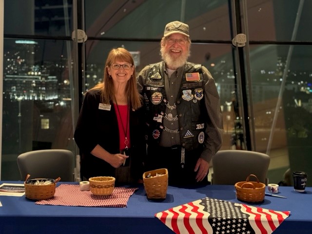 Susan, a white woman with shoulder-length brown hair and glasses in a black blazer and red blouse and Robert, a white man in a U.S. Army veteran black leather vests adorned with medallions and American flag patches, are both smiling and standing behind a table with a blue tablecloth, baskets with giveaway items, and American flag motifs. 