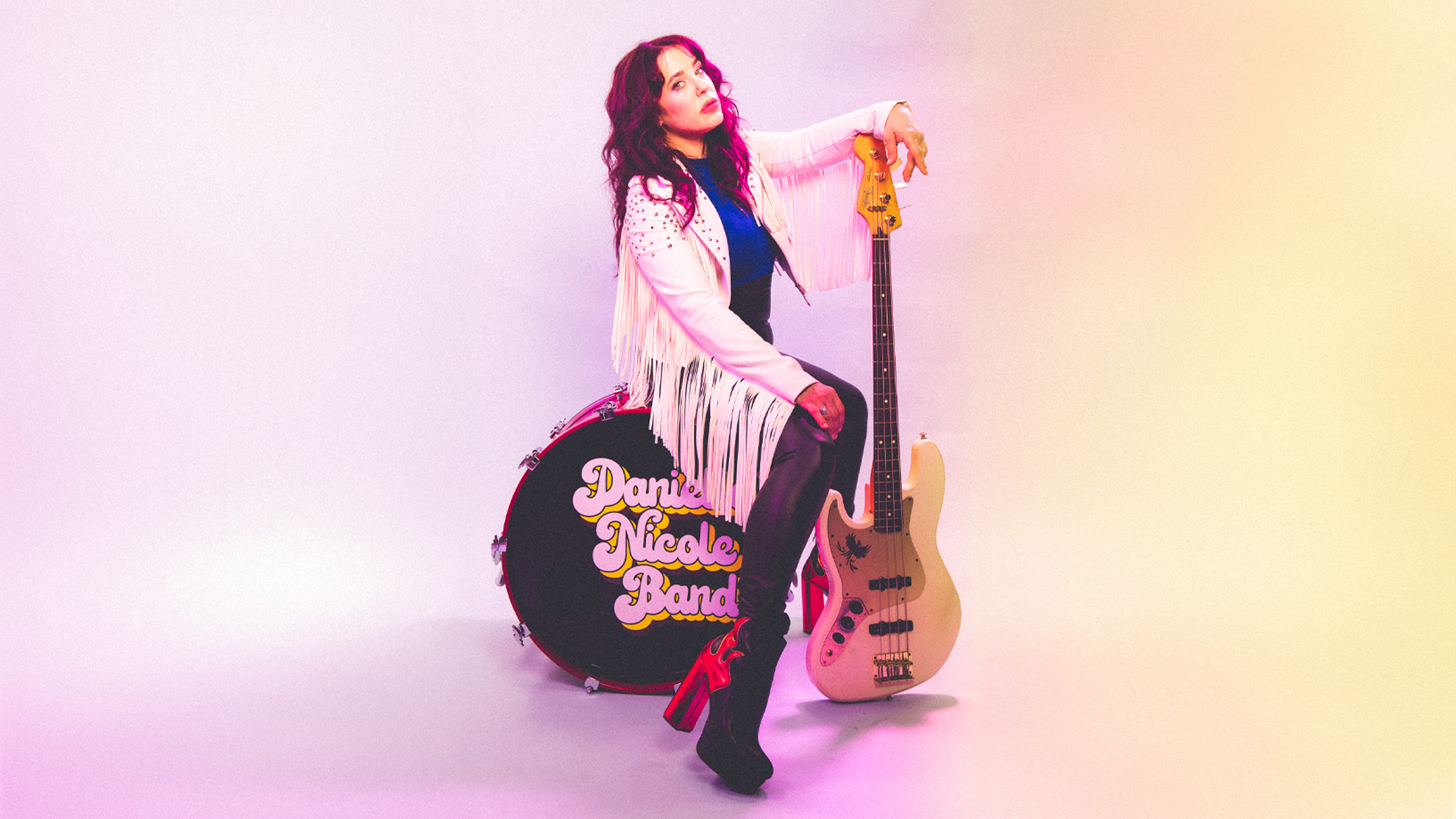 A woman with a white leather jacket sits on a bass drum that says "Danielle Nicole Band" on the side. She is leaning on a white electric guitar.